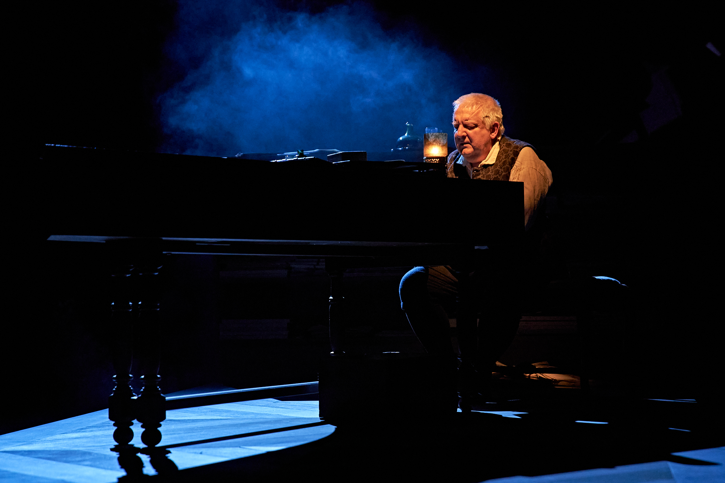 A man sits at a harpsichord in the warm glow of candlelight.