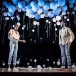 A female and male figure standing on stage with white and blue balloons floating above them.