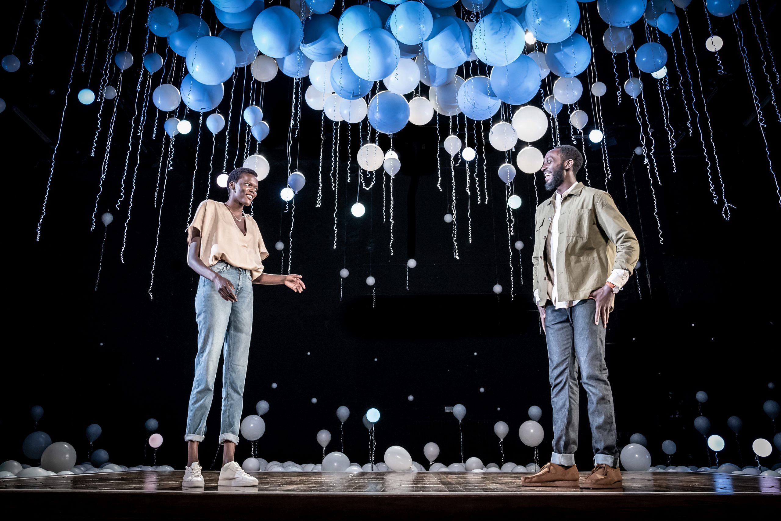 A female and male figure standing on stage with white and blue balloons floating above them.
